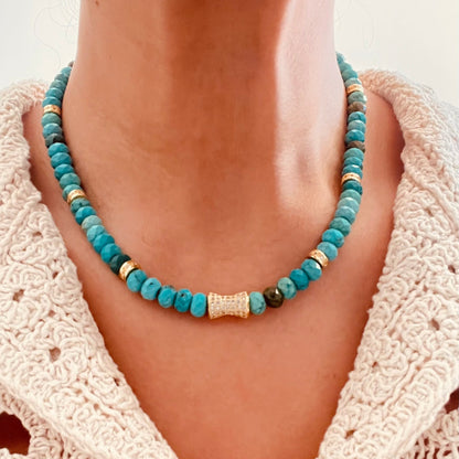 turquoise beaded necklace with a textured gold cylindrical centerpiece, paired with a cream-colored crochet knit top.